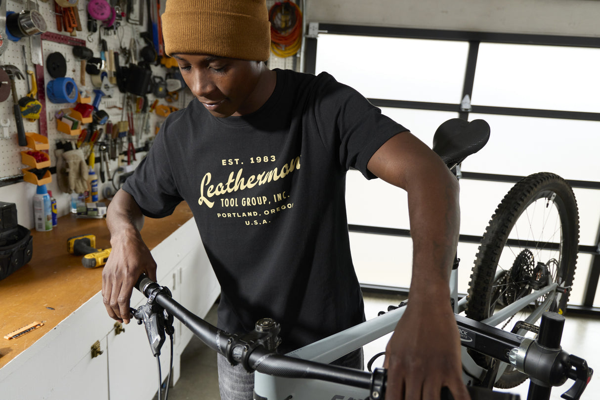Translation missing: de.A person wearing a black Leatherman Vintage Tee working on a mountain bike in a garage workshop, surrounded by tools and equipment