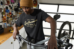 Translation missing: de.A person wearing a black Leatherman Vintage Tee working on a mountain bike in a garage workshop, surrounded by tools and equipment
