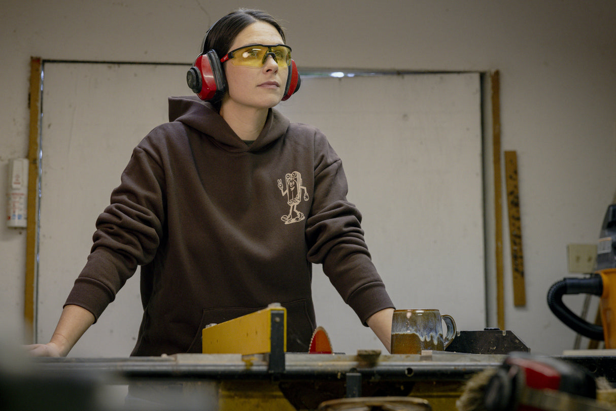 Translation missing: en.A person wearing a brown Leatherman Mr. Wave hoodie standing at a workbench in a workshop, wearing safety glasses and earmuffs