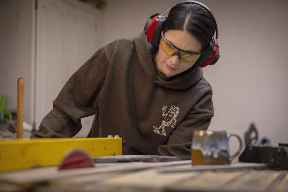 Translation missing: en.A person wearing a brown Leatherman Mr. Wave hoodie operating a table saw in a workshop, wearing safety glasses and earmuffs for protection