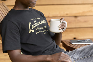 Translation missing: de.A person wearing a black Leatherman Vintage Tee relaxing on a wooden deck chair, holding a white mug with a notebook and pen on a small table nearby