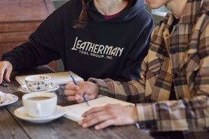Translation missing: en.Two people sitting at a table with coffee, pastries and notebooks. One is wearing a black Leatherman Blade &amp; Grain hoodie with &#39;Leatherman Tool Group Inc.&#39; on the front