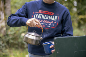 Translation missing: pl.Close-up of a person wearing a navy blue Leatherman PNW Crewneck sweatshirt while pouring hot water from a kettle into a blue enamel mug at a campsite