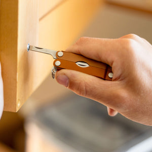 Translation missing: es.Close-up of a hand using a Pumpkin Spice Leatherman Micra multi-tool to tighten a screw on a wooden drawer, showcasing its compact design and built-in flathead screwdriver