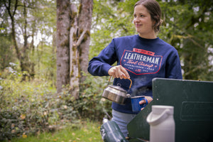 Translation missing: pl.A person wearing a navy blue Leatherman PNW Crewneck sweatshirt pouring hot water from a kettle into a mug at a campsite surrounded by trees