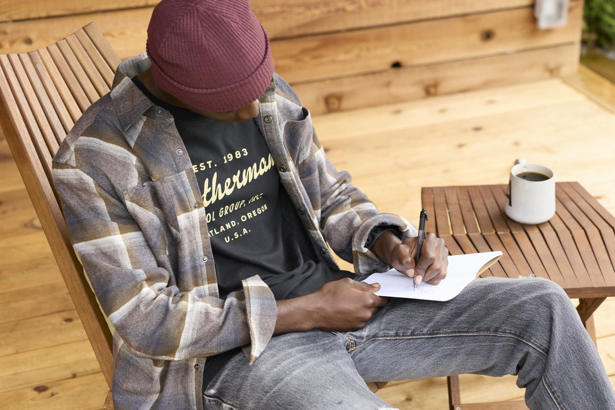 Translation missing: de.A person wearing a black Leatherman Vintage Tee sitting on a wooden chair outdoors, writing in a notebook with a mug of coffee on a nearby table