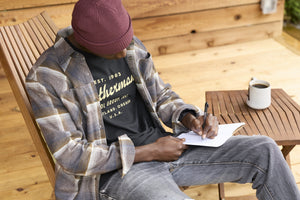 Translation missing: de.A person wearing a black Leatherman Vintage Tee sitting on a wooden chair outdoors, writing in a notebook with a mug of coffee on a nearby table