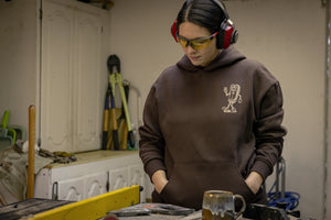 Translation missing: en.A person wearing a brown Leatherman Mr. Wave hoodie standing in a workshop with safety glasses and ear protection, looking down at a workbench with tools and a ceramic mug