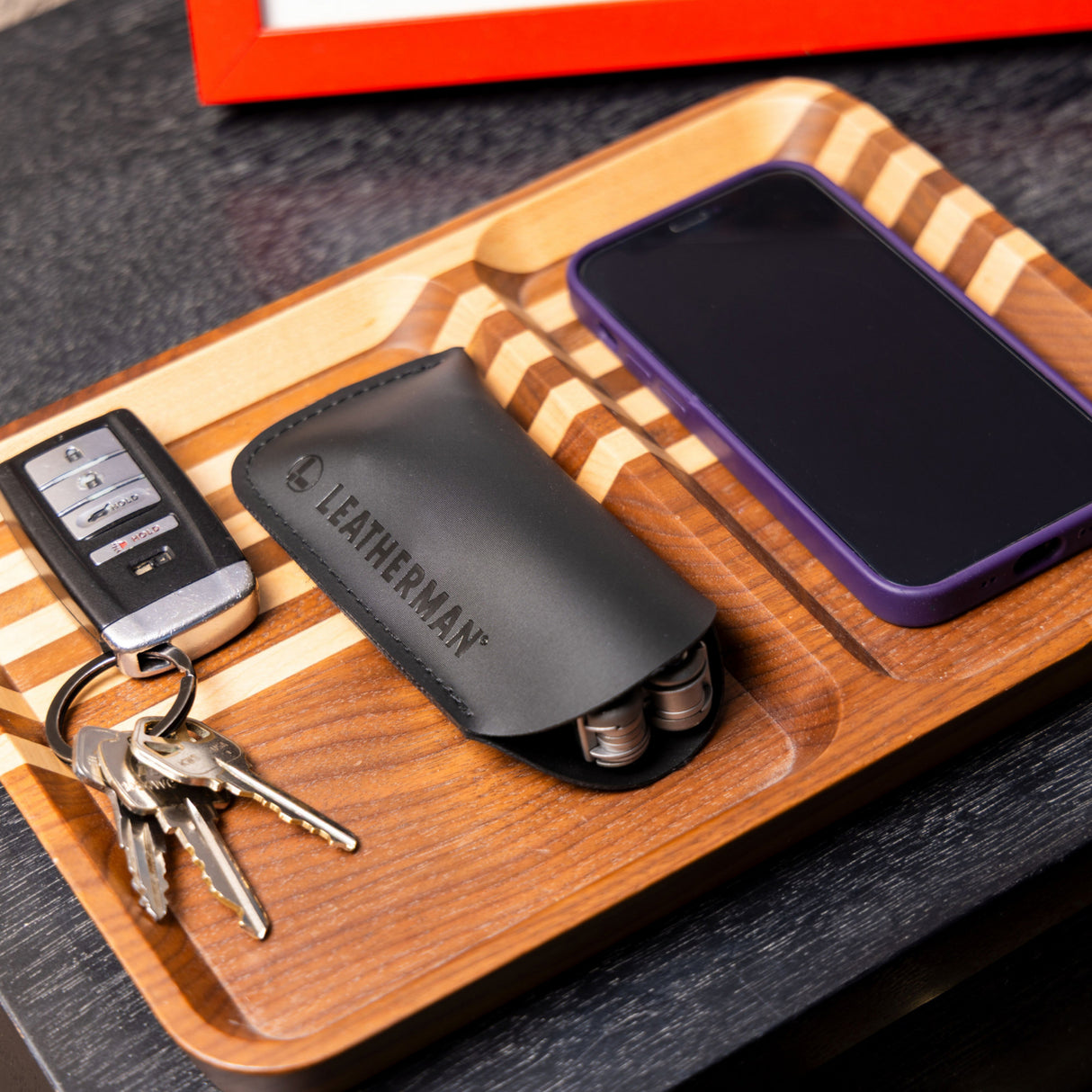 Translation missing: es.A black Leatherman Hypalon tool sleeve holding a multi-tool, neatly placed on a wooden valet tray beside a set of keys and a smartphone with a purple case, showcasing an organized everyday carry setup