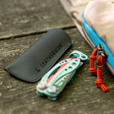 Translation missing: es.A black Leatherman Hypalon tool sleeve placed on a wooden surface next to a folded Paradise-colored Skeletool® CX multi-tool, with a beige and blue-zippered pouch partially visible in the background