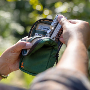 Translation missing: de.A person opens a Leatherman Tool Pouch in Hunter color, retrieving a Leatherman Arc® from the organized interior while outdoors, with sunlight filtering through blurred green foliage in the background