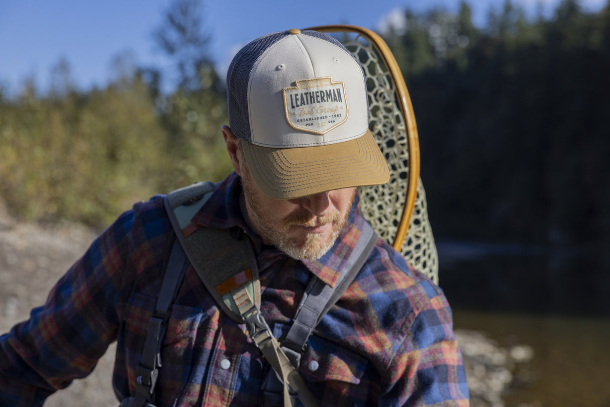Translation missing: es.A person wearing a tan and brown Leatherman PNW Trucker Hat standing near a river with a backpack and fishing net