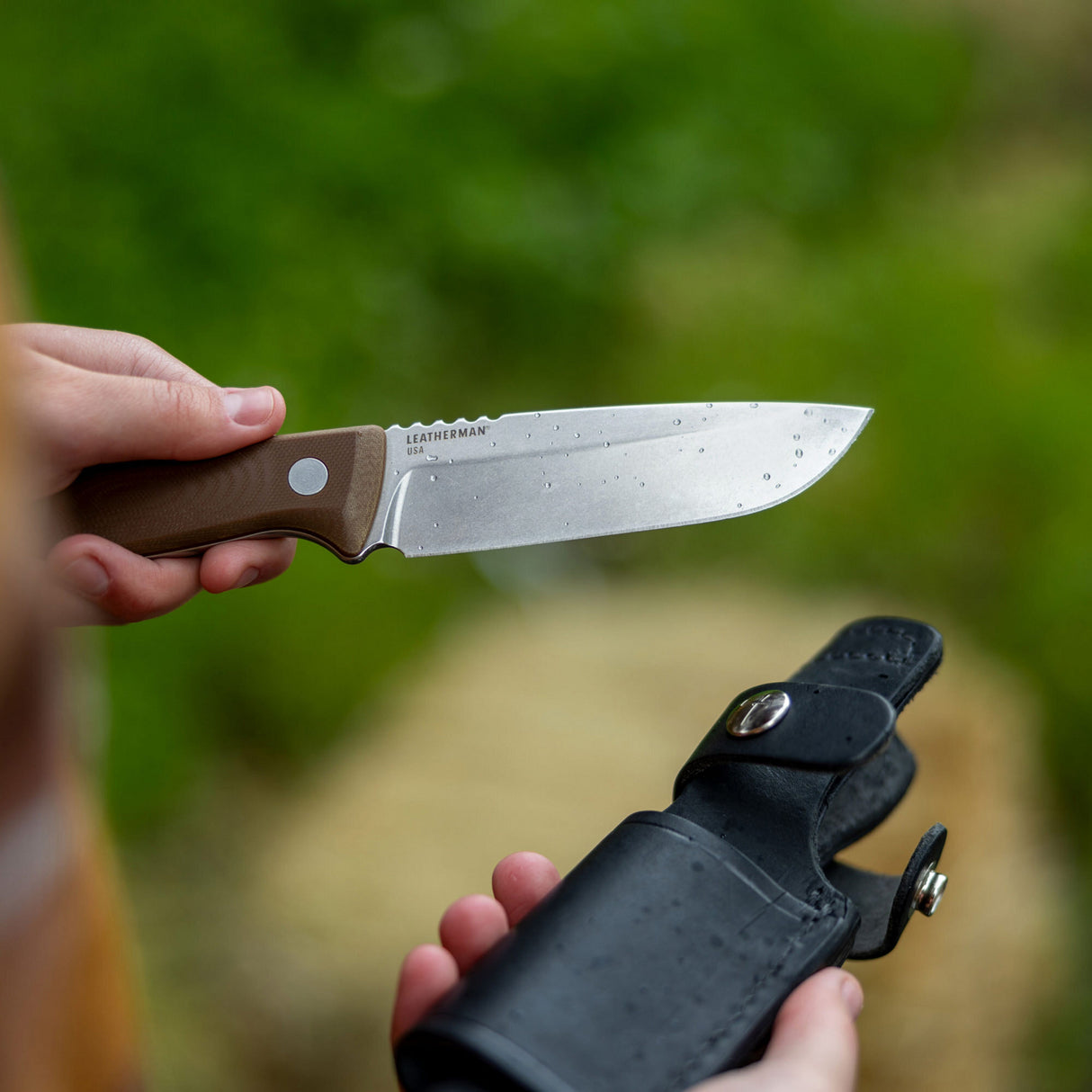 Translation missing: fr.Person holding a Leatherman Trac Earth Brown fixed-blade knife with water droplets on the blade, positioned above a black leather sheath in an outdoor setting with green foliage in the background