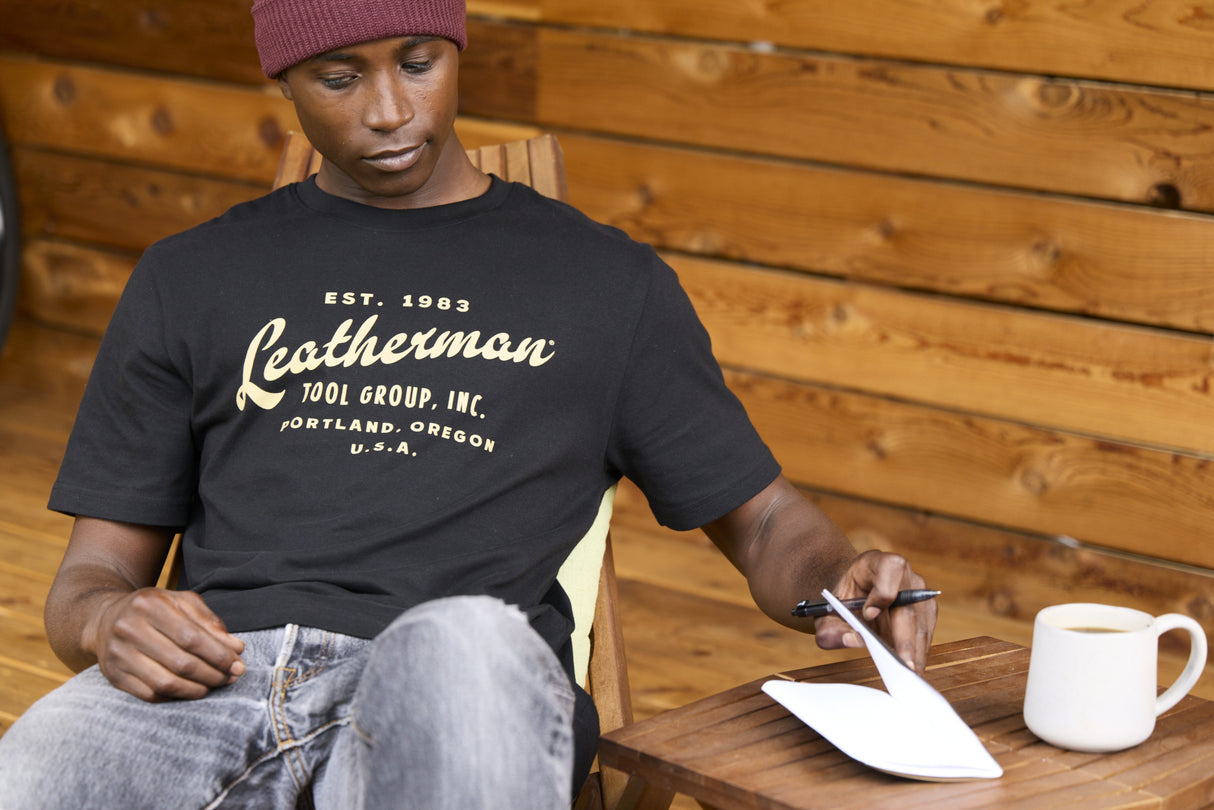 Translation missing: de.A person wearing a black Leatherman Vintage Tee sitting on a wooden deck chair, opening a notebook beside a mug of coffee