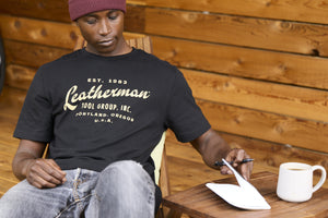 Translation missing: de.A person wearing a black Leatherman Vintage Tee sitting on a wooden deck chair, opening a notebook beside a mug of coffee