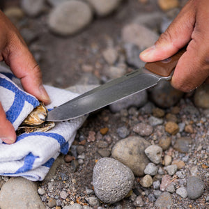 Translation missing: fr.Person using a Leatherman Trac Earth Brown fixed-blade knife to open a shellfish wrapped in a towel on a rocky surface, highlighting outdoor food preparation