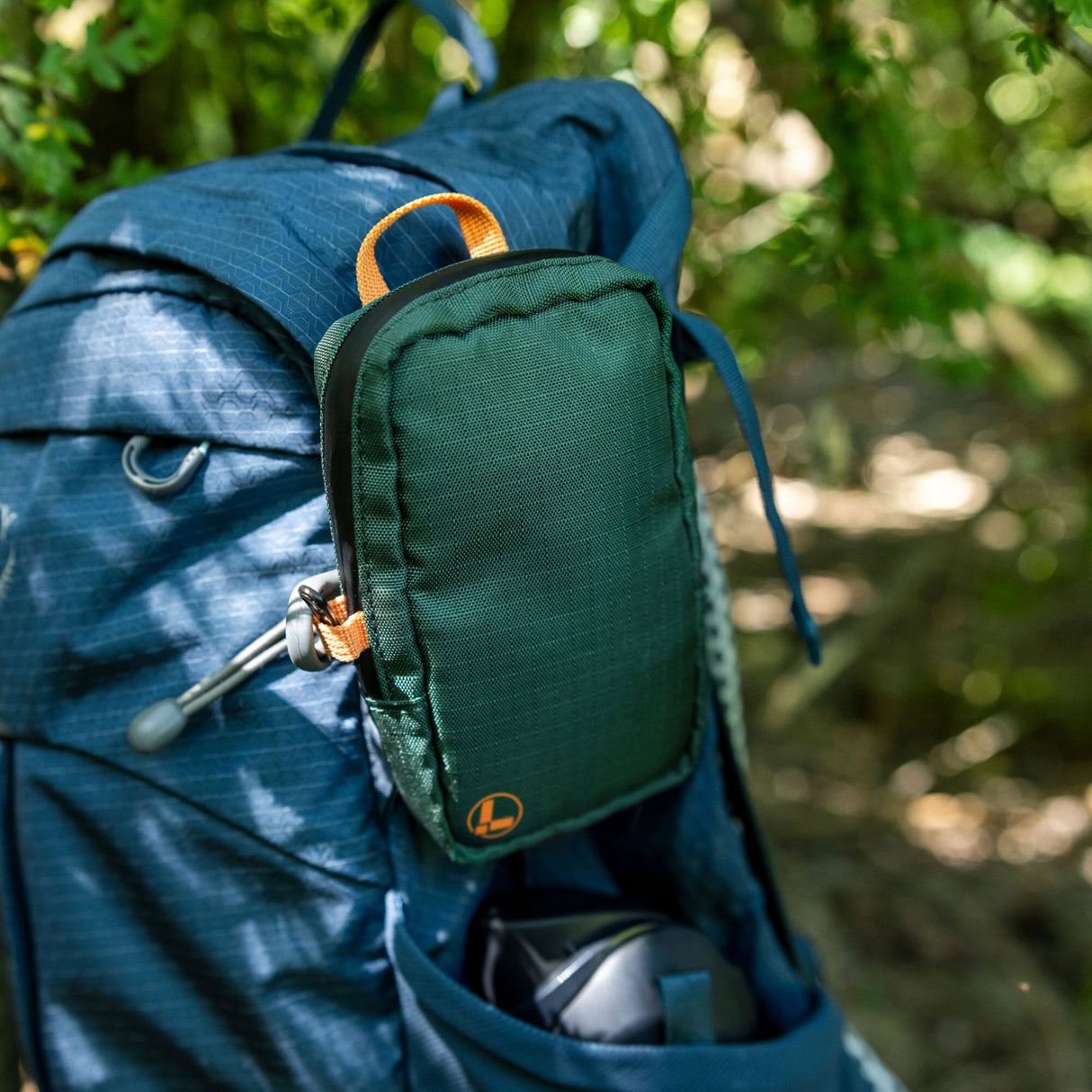 Translation missing: de.A Leatherman Tool Pouch in Hunter color is clipped to the shoulder strap of a blue hiking backpack, resting in an outdoor forest setting with sunlight filtering through the trees