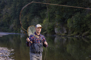 Translation missing: es.A person wearing a beige Leatherman PNW Trucker hat fly-fishing in a river surrounded by trees