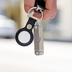 Translation missing: es.A Winter Fog colored Leatherman Micra multi-tool attached to a keyring alongside a black Apple AirTag holder, held by a person's hand