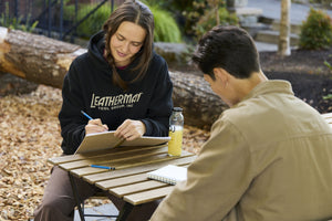 Translation missing: en.Two people sitting at a table outside with notebooks and pencils. One is wearing a black Leatherman Blade &amp; Grain Hoodie with &#39;Leatherman Tool Group Inc.&#39; on the front