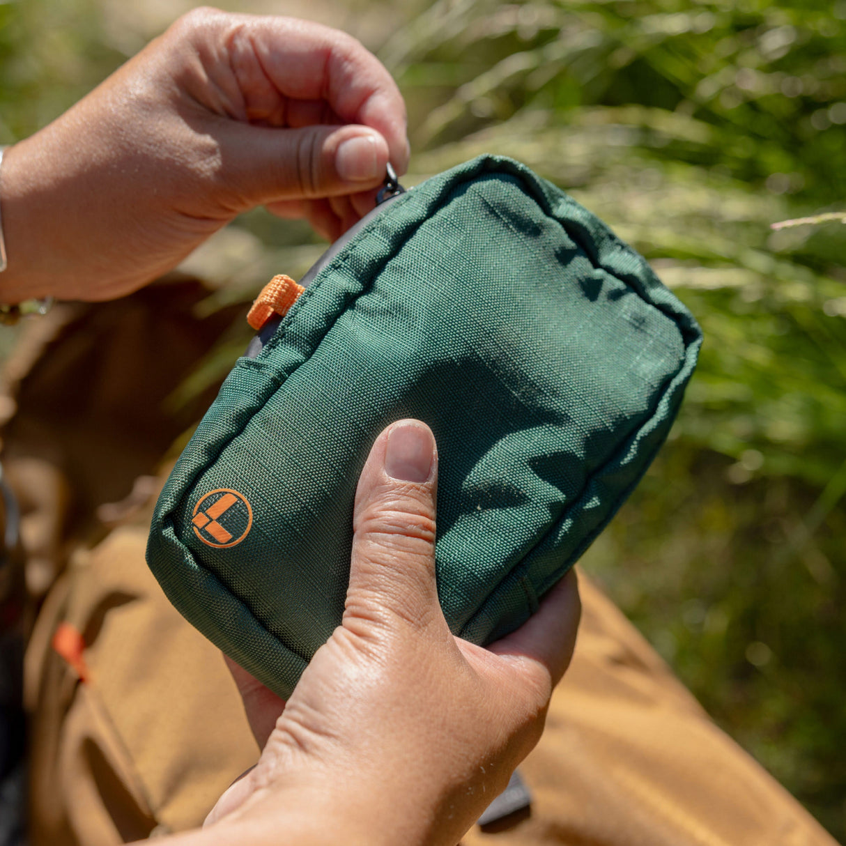 Translation missing: de.A person holds and unzips a Leatherman Tool Pouch in Hunter color outdoors, featuring a deep green fabric with an orange logo and tab, set against a background of grass and a brown bag