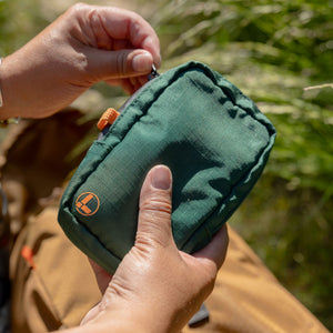 Translation missing: de.A person holds and unzips a Leatherman Tool Pouch in Hunter color outdoors, featuring a deep green fabric with an orange logo and tab, set against a background of grass and a brown bag