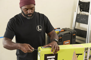 Translation missing: es.A person wearing a black Leatherman Classic Tee using the blade on a Wave Alpha in Cascadia color to open a large box in a workshop while standing near a ladder and tool case
