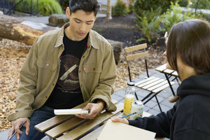 Translation missing: es.A man sitting at a table wearing a black Leatherman Blade &amp; Grain tee with a woman wearing a black hoodie, both equipped with a notebook and pencil