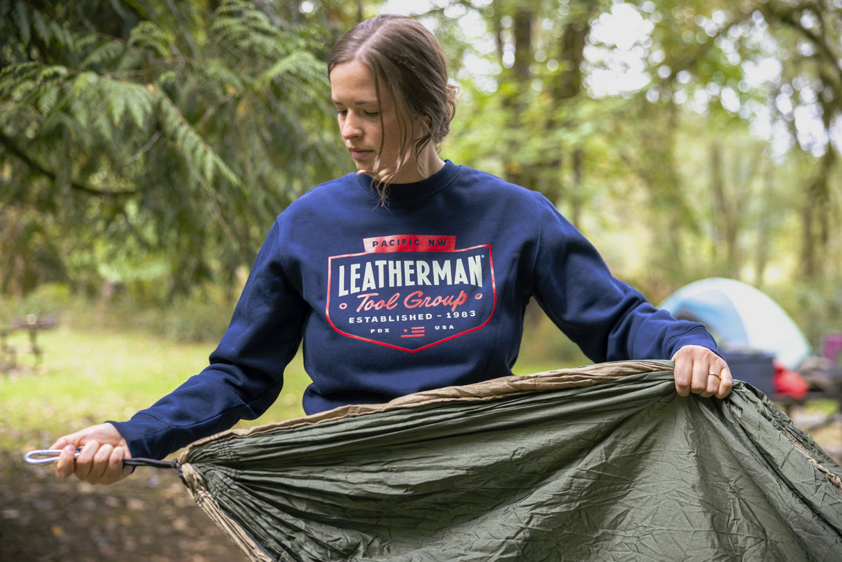 Translation missing: pl.A person wearing a navy blue Leatherman PNW Crewneck sweatshirt setting up a hammock at a forest campsite with a tent in the background