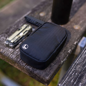 Translation missing: en.A black Leatherman Tool Pouch with a white logo rests on a weathered wooden bench outdoors, positioned beside a Leatherman ARC multi-tool and black water bottle, suggesting a rugged, ready-for-anything everyday carry setup