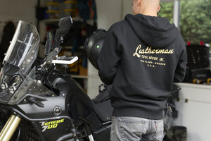 Translation missing: en.A person wearing a black Leatherman Vintage Zip-Up Hoodie standing next to a motorcycle, showing the gold Leatherman Tool Group, Inc. logo printed on the back