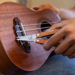 Translation missing: es.A hand uses a Leatherman Micra multi-tool in Pumpkin Spice color to trim string ends on a wooden ukulele, showcasing the tool's compact scissors in action