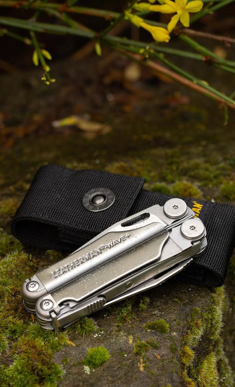 A Leatherman Wave with dew on it laying on a mossy rock on top of a Leatherman black nylon sheath.