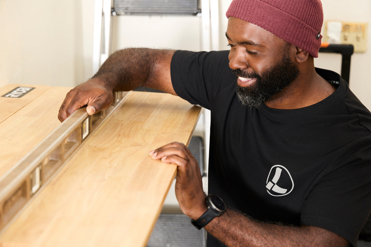Translation missing: en.A person wearing a black Leatherman Classic Tee using a level tool to level a wooden shelf indoors