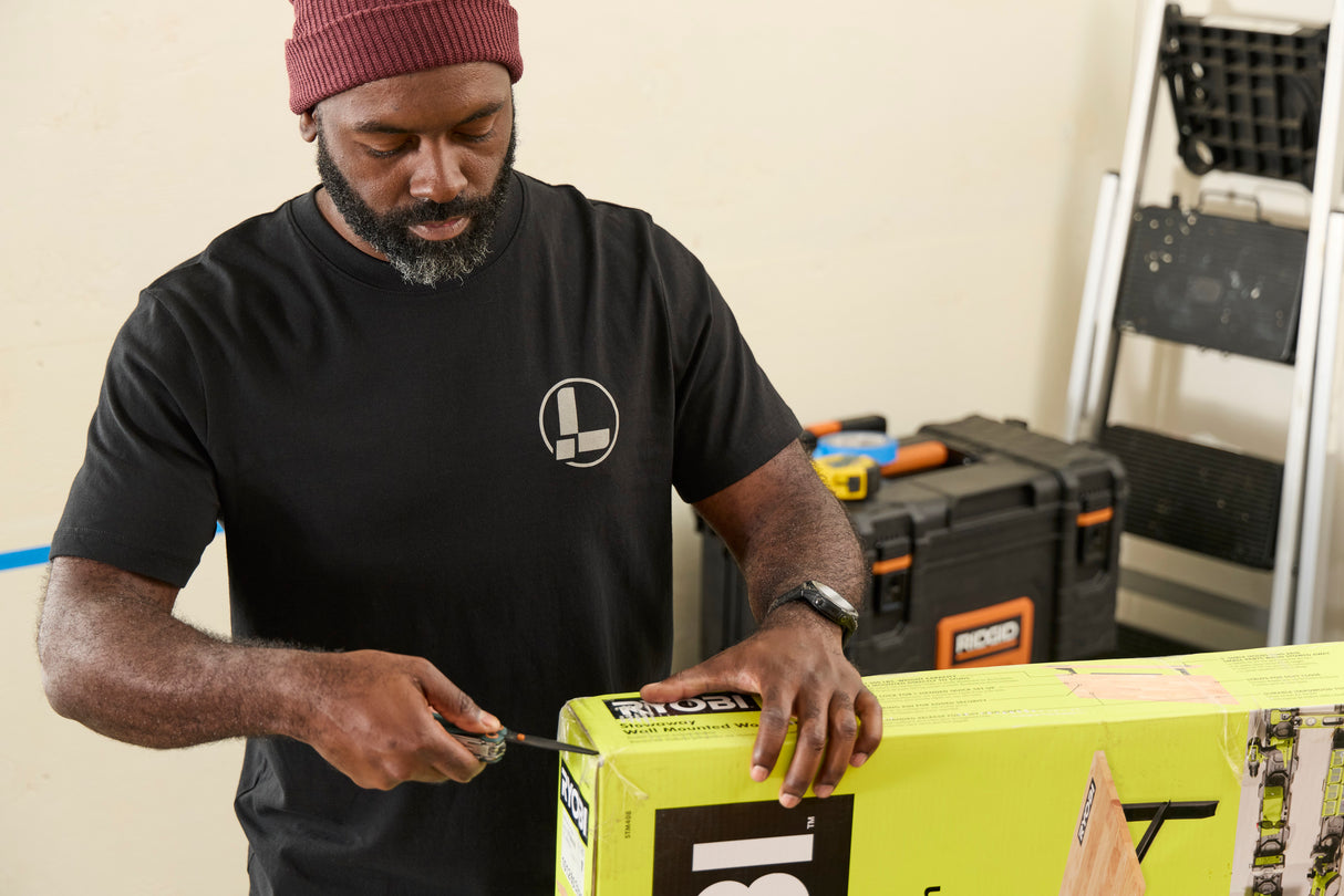Translation missing: en.A person wearing a black Leatherman Classic Tee using the blade on a Wave Alpha in Cascadia color to open a large box in a workshop while standing near a ladder and tool case
