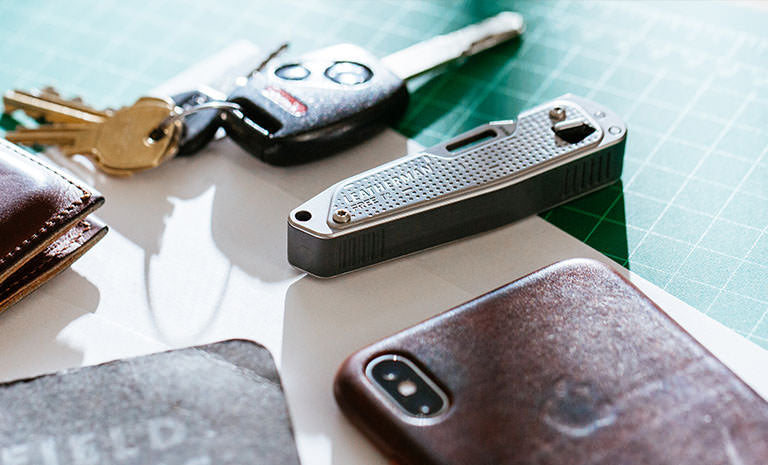 Translation missing: en.Brown Leatherman Wallet, car keys, Leatherman T2, gray note pad, and Iphone on top of white piece of paper placed on green grid table