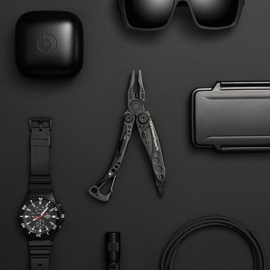 A collection of accessories including a black watch, sunglasses, earphones, and Leatherman Skeletool Cx displayed on a sleek black surface.