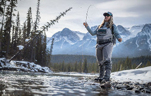 Translation missing: nl.A woman stands in shallow water, casting a fly rod, surrounded by nature and reflecting a peaceful fishing scene