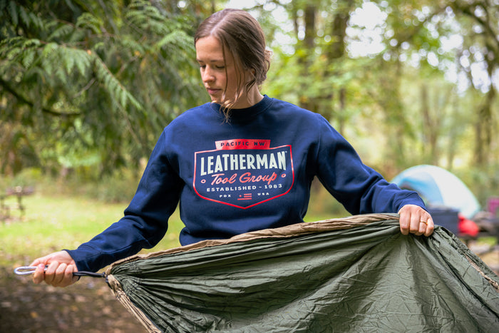 Translation missing: de.A woman wearing a navy Leatherman Pacific Northwest Crewneck holding a hammock in a forest setting