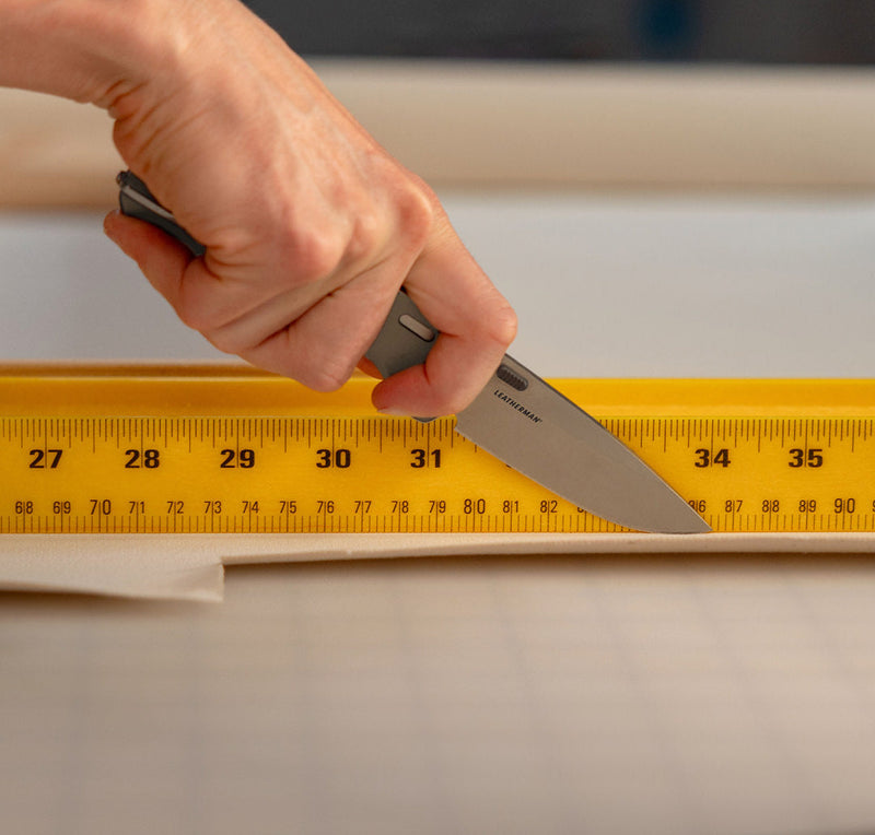 Close-up image of a hand using a Leatherman folding knife to cut fabric along a yellow measuring ruler on a gridded cutting mat, illustrating precision and utility in a workspace or crafting environment.