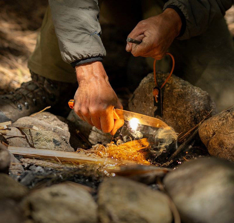 Person igniting a fire using a ferro rod and a Leatherman fixed-blade knife with an orange handle, creating sparks over kindling in a rocky outdoor campsite setting.