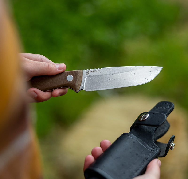 A person holding a Leatherman fixed-blade knife with a brown handle and a wet blade, positioned above a black leather sheath in the other hand. The background features soft-focus greenery, suggesting an outdoor or wilderness setting.