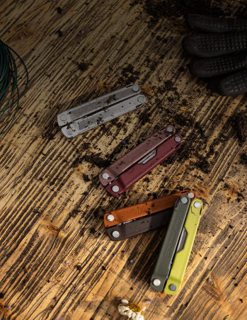 Four differently colored Leatherman Bond tools closed on a wooden floor with dirt sprinkled on them.