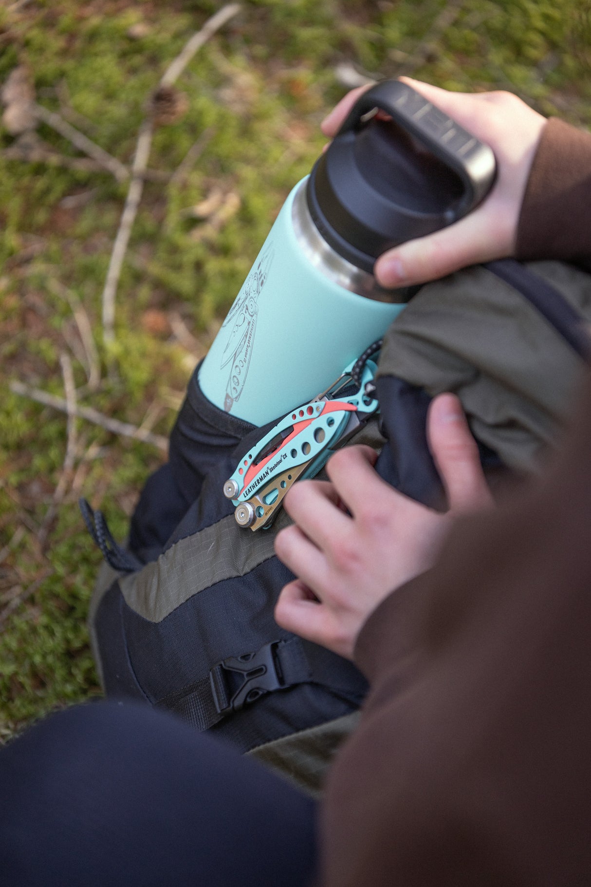 Translation missing: pl.Hand holding a mint green Yeti bottle in a backpack pocket with a Paradise Leatherman Skeletool CX multitool clipped on