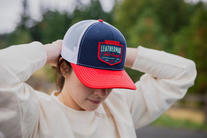 Translation missing: en.A person adjusting a navy Leatherman PNW Trucker Hat with a white mesh back and red brim while outdoors