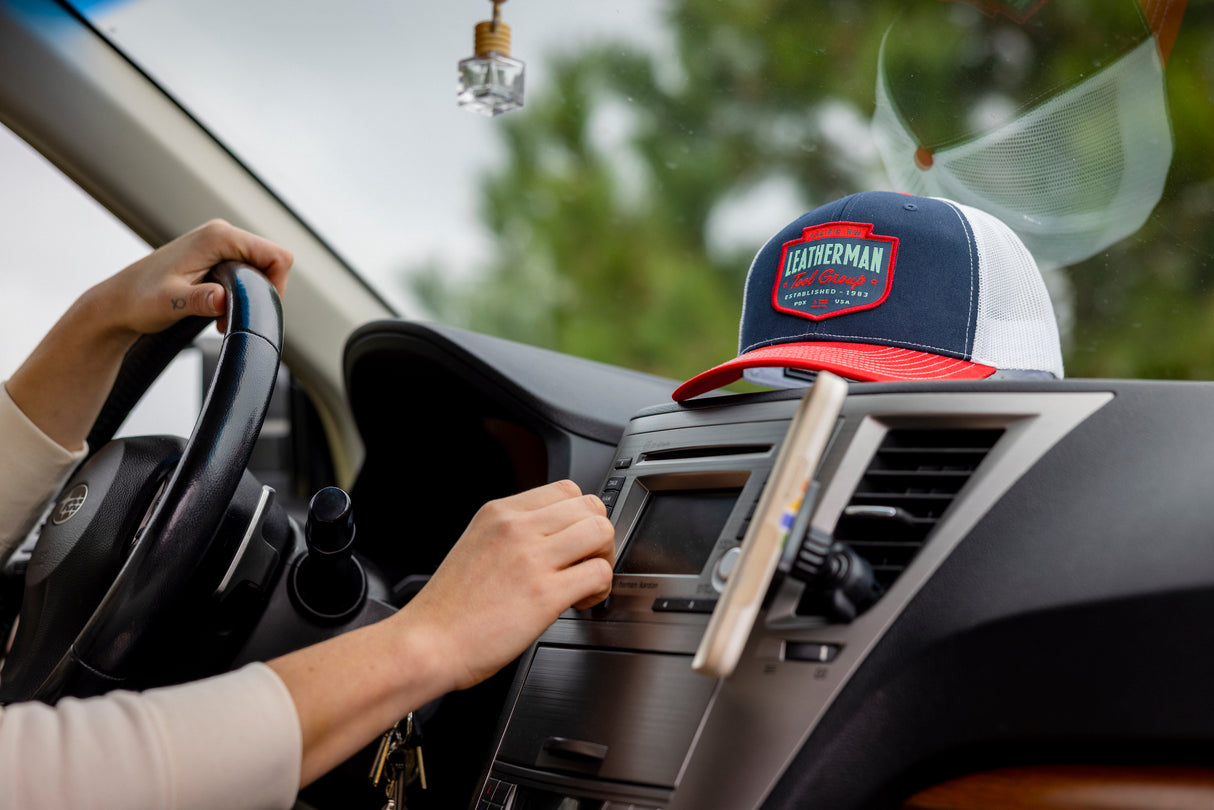 Translation missing: en.A person in a car with a navy Leatherman PNW Trucker Hat with a white mesh back and red brim resting on the dashboard