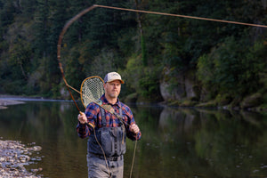 Translation missing: de.A person wearing a beige Leatherman PNW Trucker hat fly-fishing in a river surrounded by trees