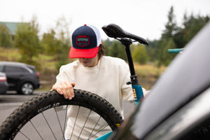 Translation missing: en.A person wearing a navy Leatherman PNW Trucker Hat with a white mesh back and red brim loading a blue mountain bike onto a vehicle