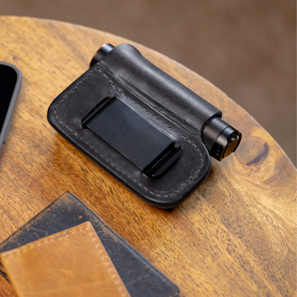 Translation missing: fr.Black leather sheath with belt clip holding a small cylindrical flashlight on a wooden surface