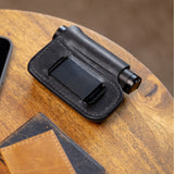 Translation missing: fr.Black leather sheath with belt clip holding a small cylindrical flashlight on a wooden surface